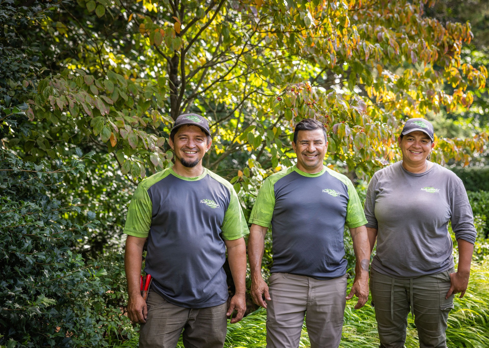 Three people stand smiling outdoors, wearing work uniforms with green accents, surrounded by lush greenery and plants in a garden setting.