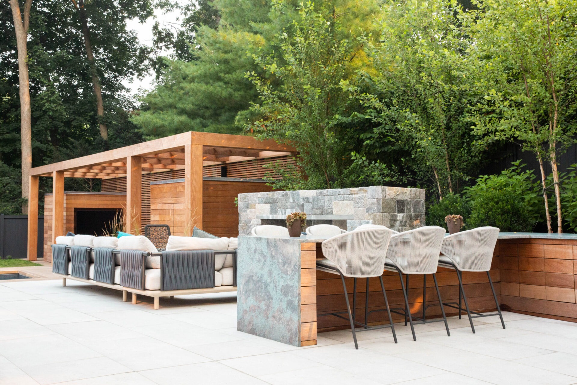Outdoor patio with modern furniture, wooden pergola, stone bar, and surrounding greenery, blending comfort with nature for relaxation and entertainment.