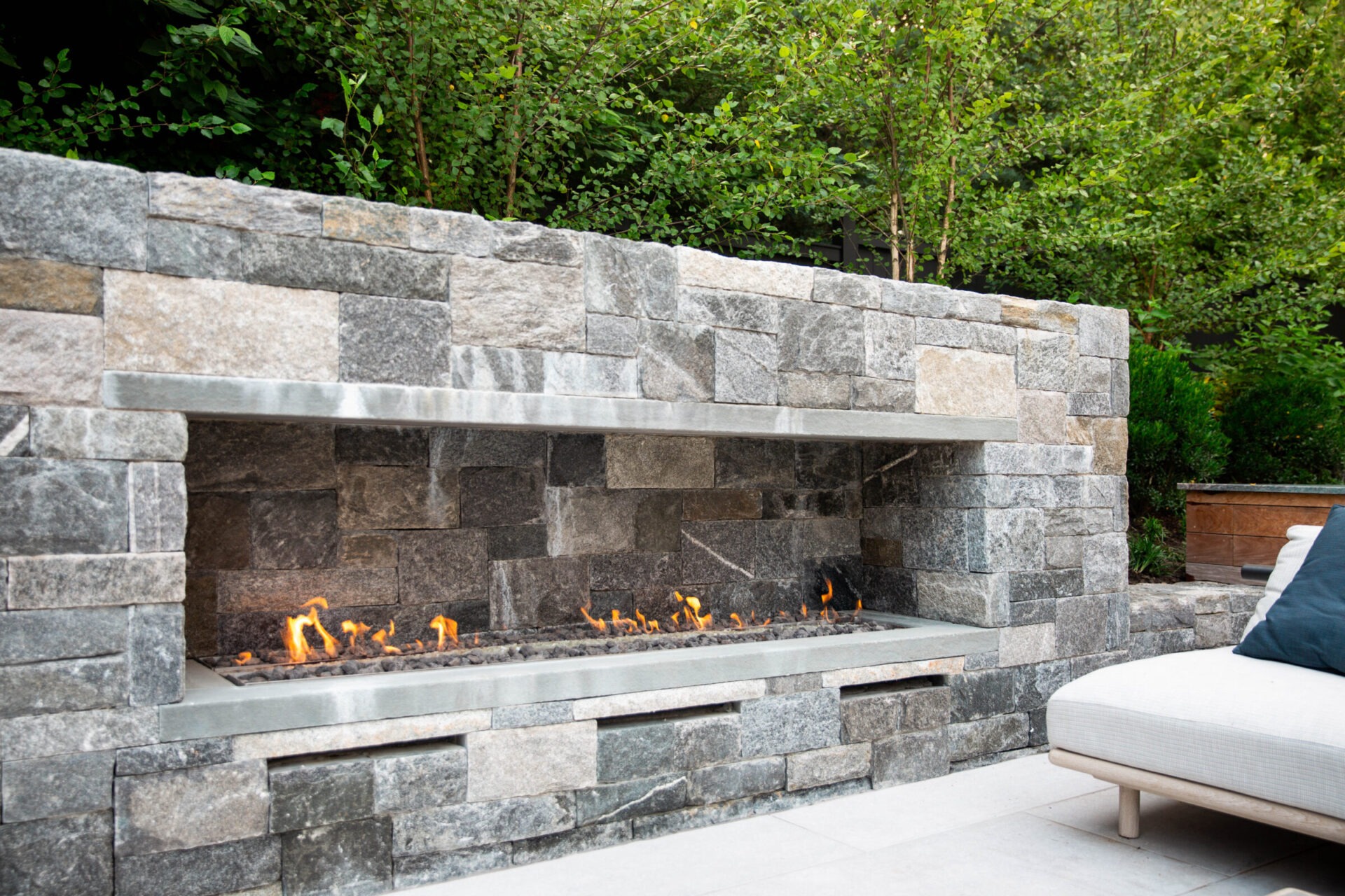 An outdoor gas fireplace with a stone surround. Flames burn over a metal ledge. A cushioned outdoor chaise lounge sits nearby, surrounded by greenery.