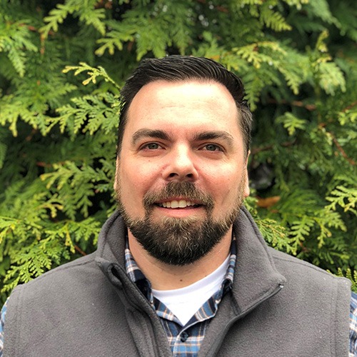 A smiling person with dark hair and beard, wearing a gray vest over a checkered shirt, stands in front of green foliage.