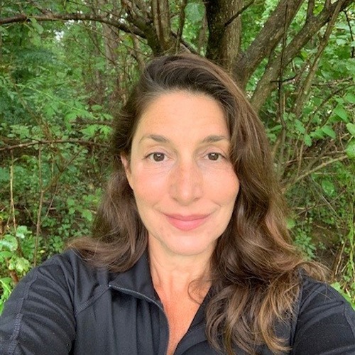 A person with long hair is smiling at the camera. They are outdoors with green foliage in the background. They're wearing a black top.