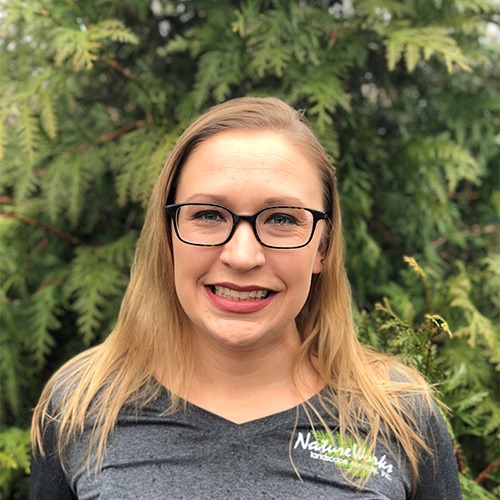 A smiling person with glasses, long straight hair, and a grey top stands before a background of lush green foliage.