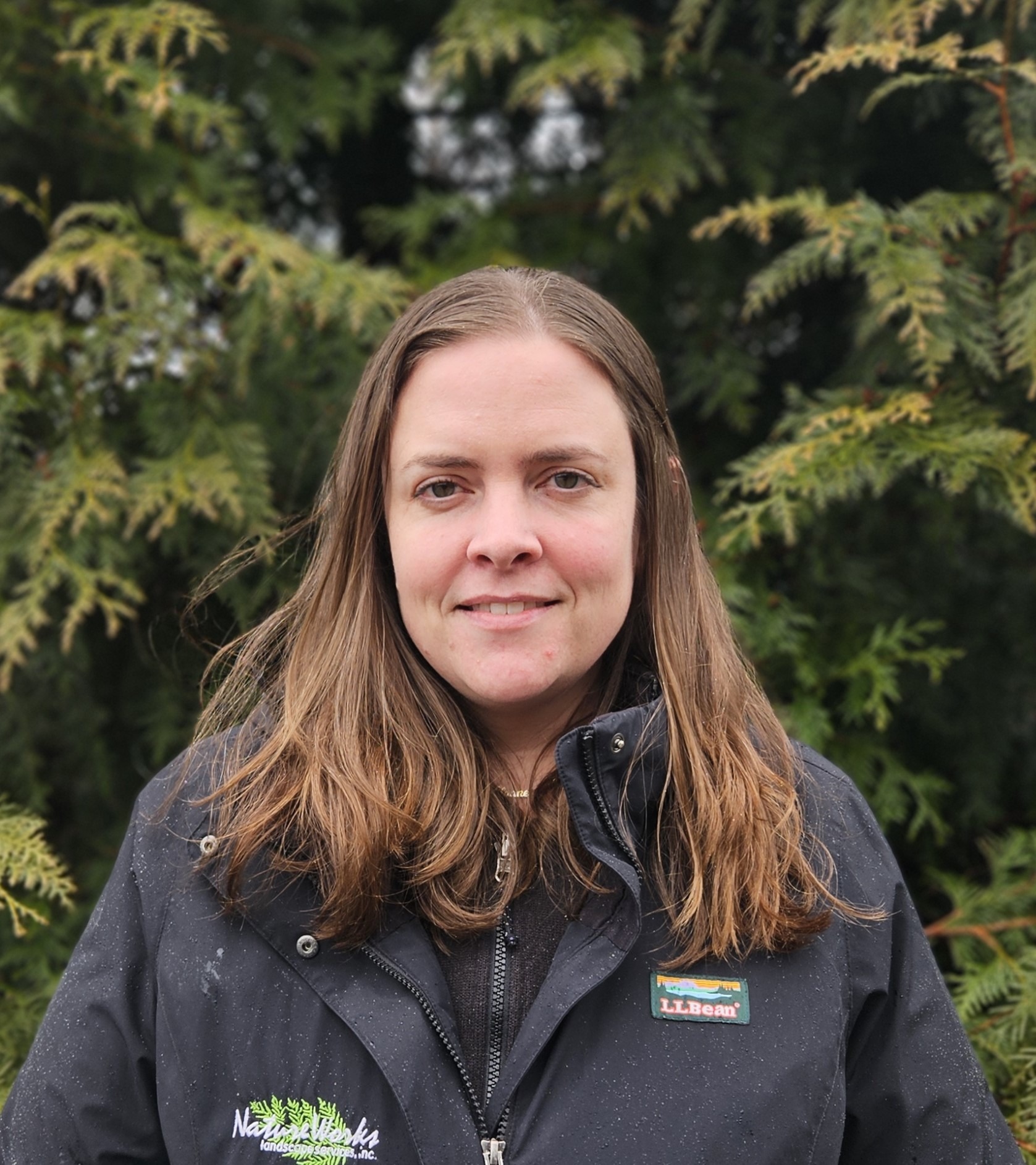 A person is standing in front of evergreen trees, wearing a black jacket with visible logos. The person has long hair and a slight smile.