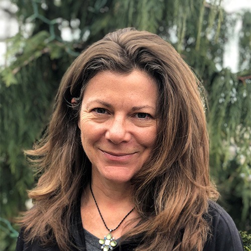 A person with shoulder-length brown hair smiling at the camera, wearing a dark top and a pendant necklace, with greenery blurred in the background.