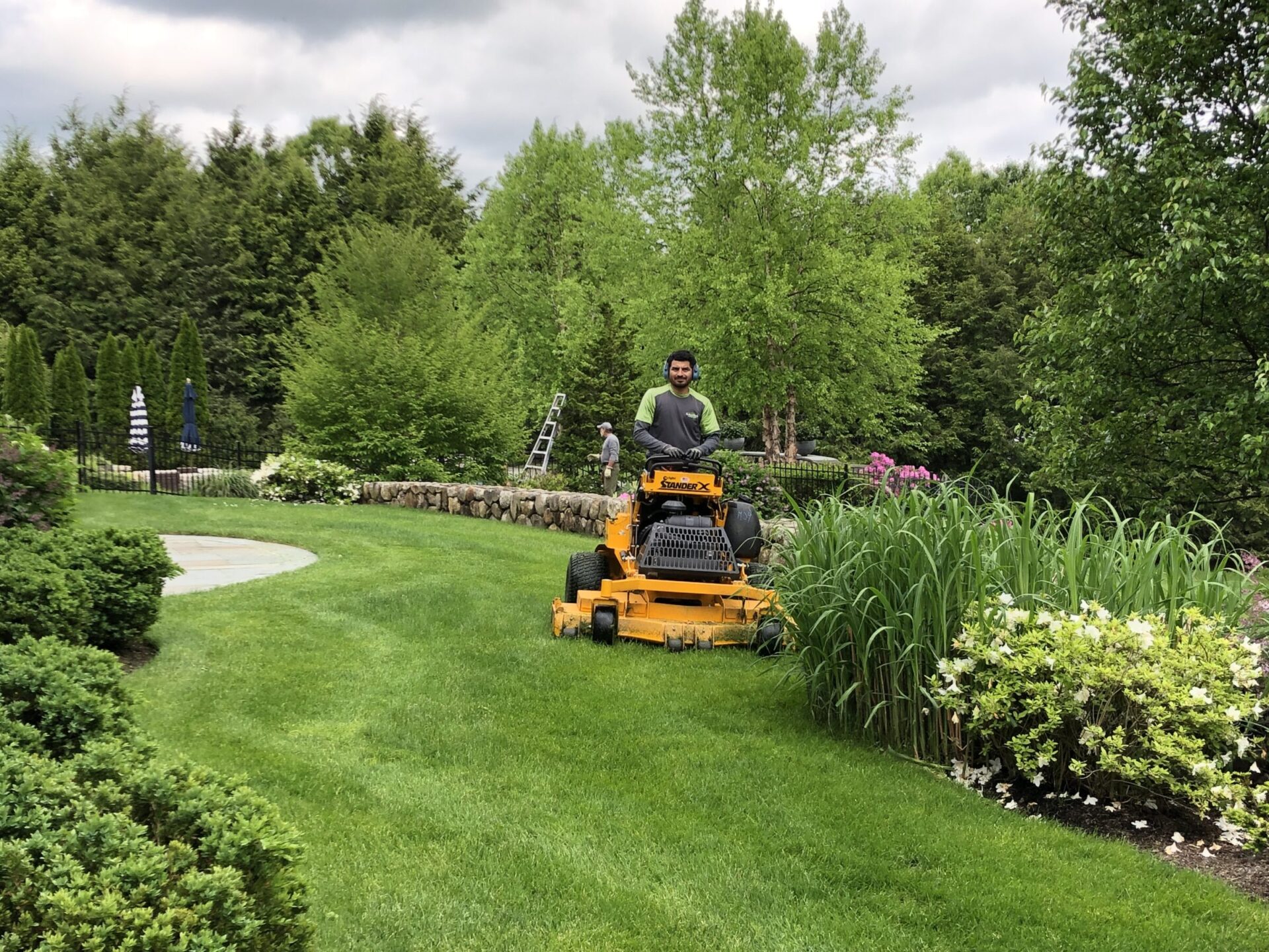 A person is riding a lawn mower in a well-manicured garden with lush greenery, flowers, and a second person standing by a ladder.