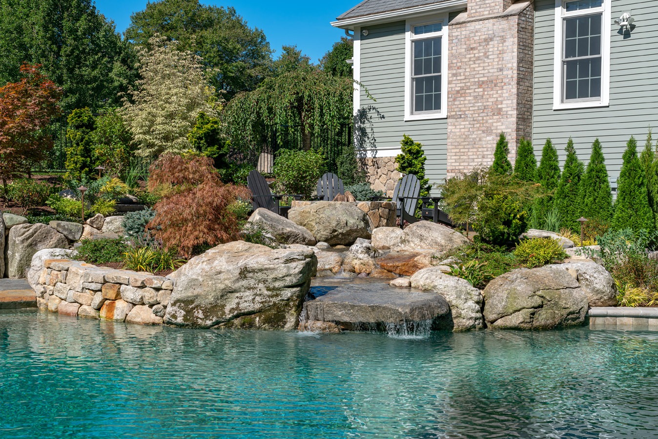 A backyard with a naturalistic swimming pool, waterfall, and landscaping, featuring rocks and diverse plants beside a house with Adirondack chairs.