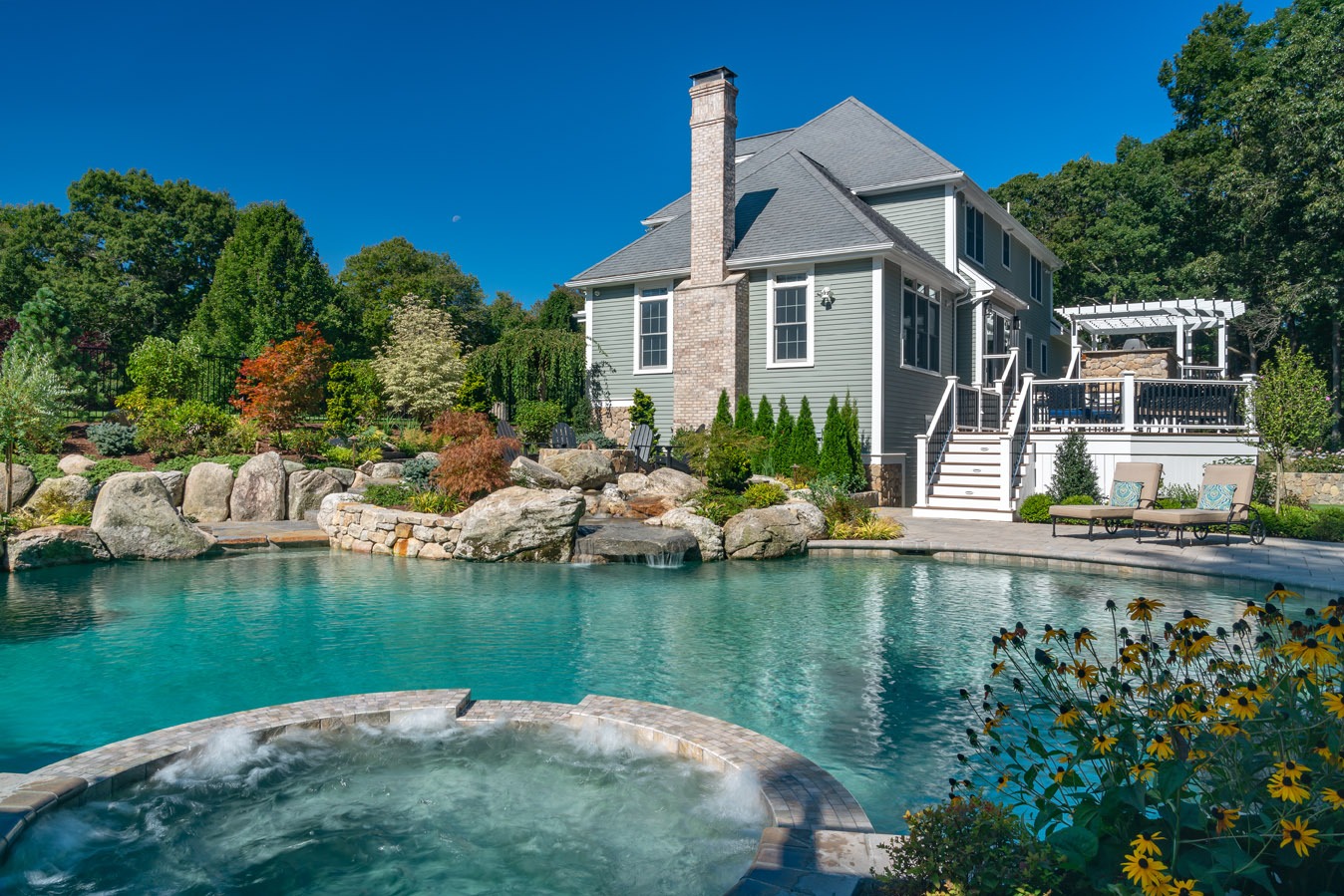 This image shows a luxurious backyard with a landscaped garden, an outdoor pool, whirlpool, and a two-story house with a spacious deck.