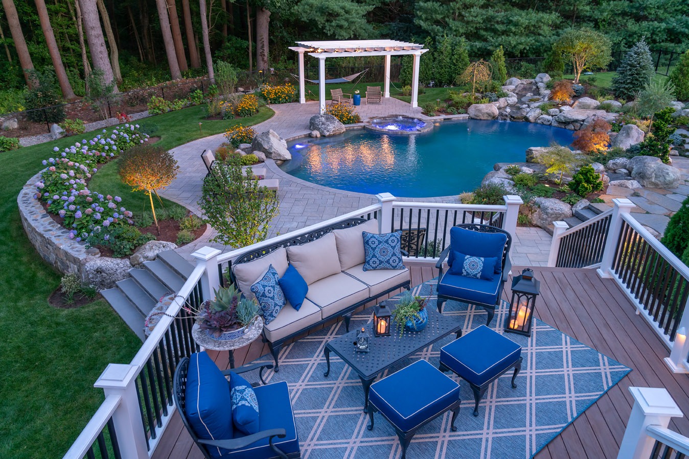 A luxurious backyard with a well-lit, curved pool, pergola, landscaped garden, and a deck with comfortable outdoor furniture and decorative cushions.