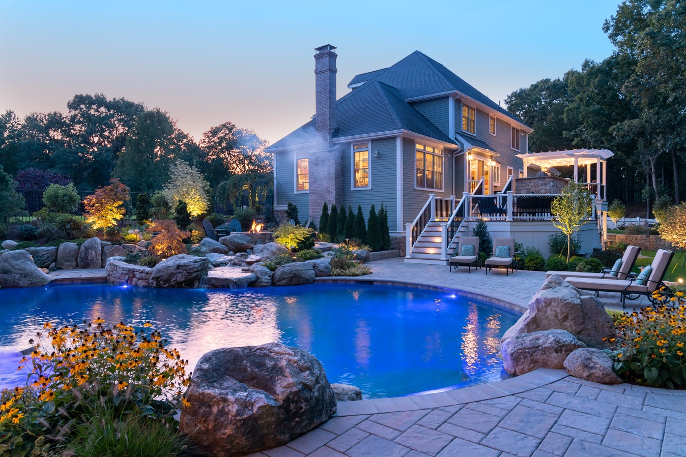 A luxurious house at dusk with lights on, featuring an illuminated blue pool surrounded by rocks, landscaping, and an inviting outdoor living space.