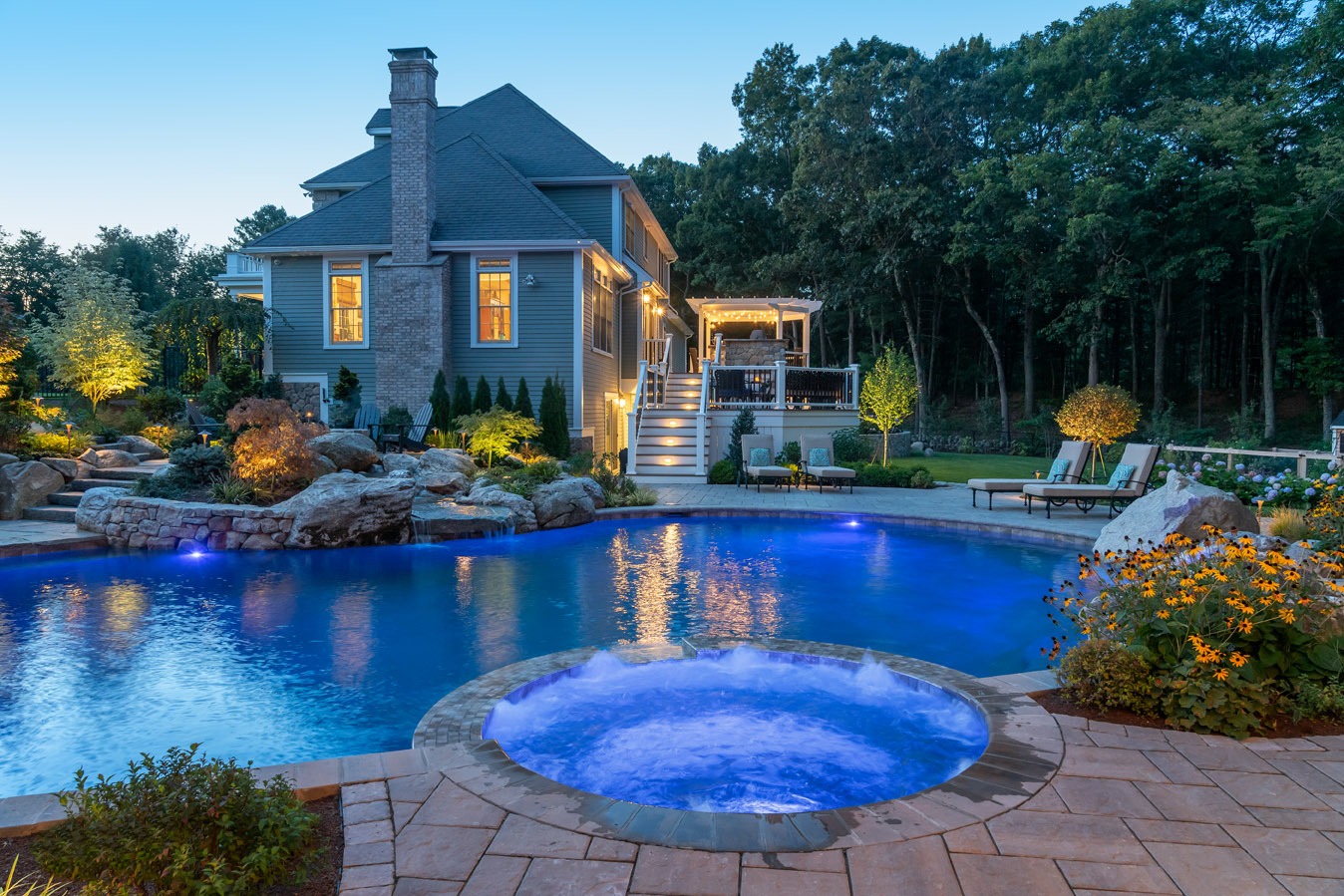 An illuminated backyard with an in-ground pool and hot tub at dusk. A luxurious house, landscaped garden, and outdoor furniture are visible.