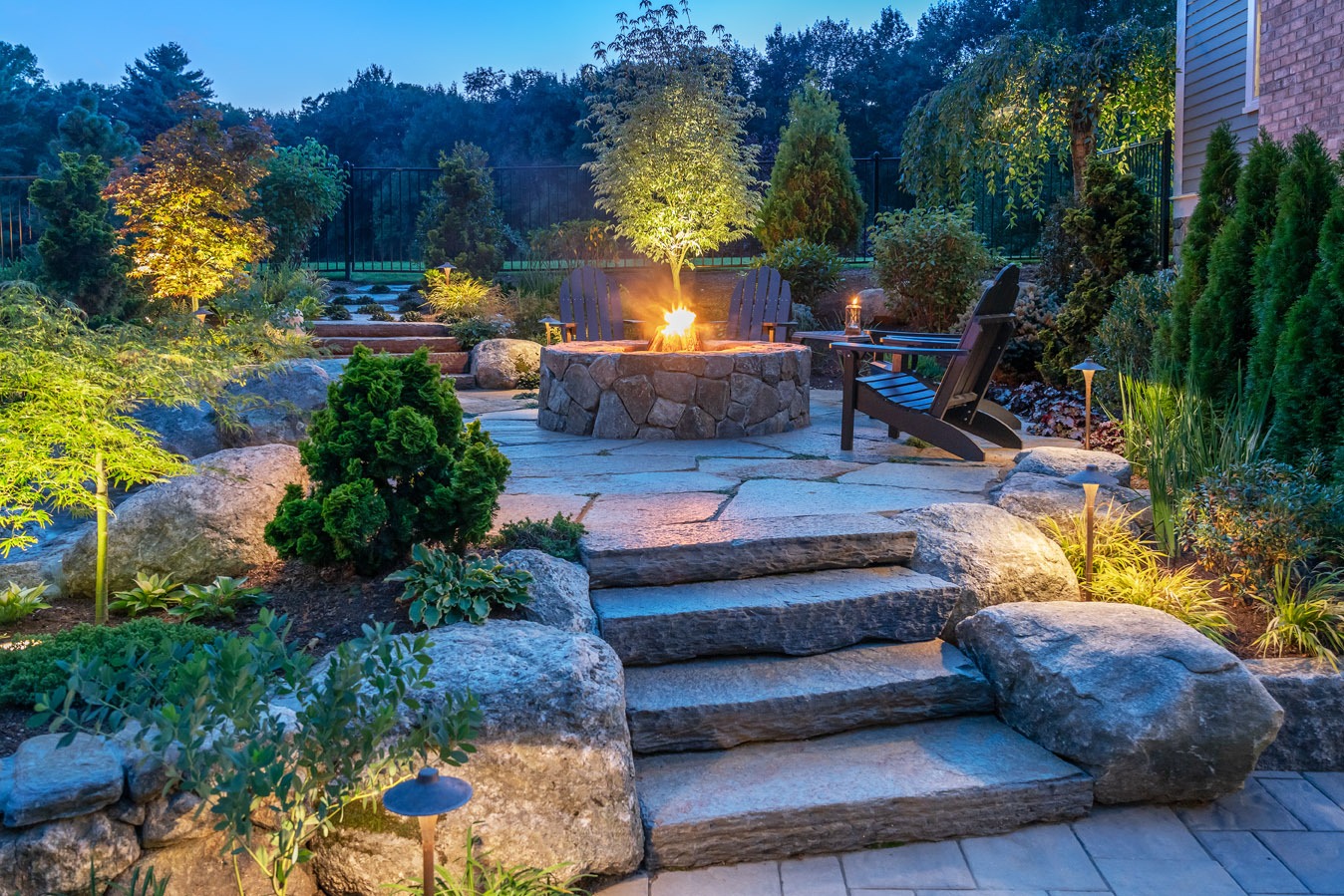 An inviting outdoor space at twilight featuring stone steps, a fire pit, Adirondack chairs, and illuminated landscaping creating a peaceful evening ambiance.