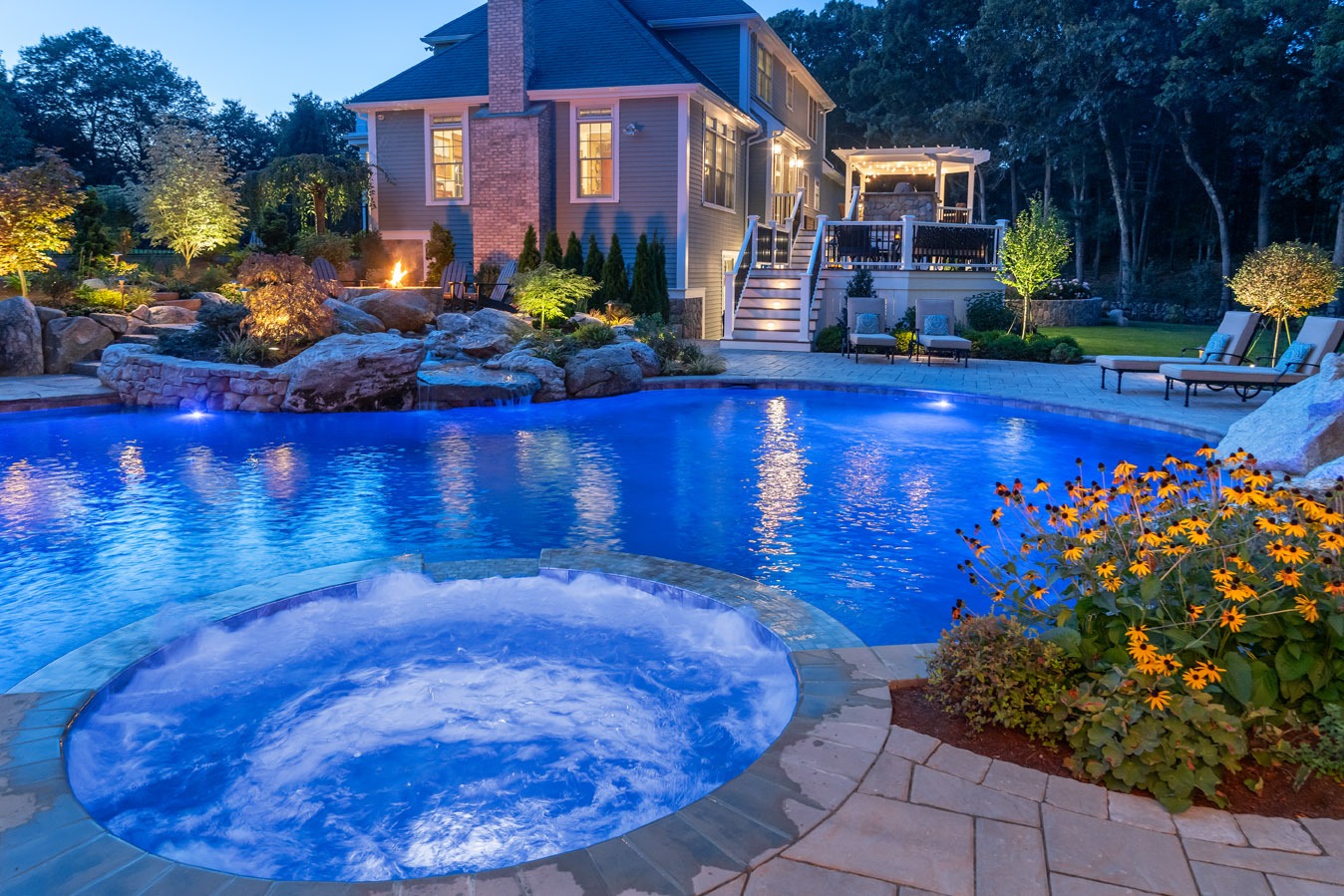 Luxurious backyard with illuminated inground pool and jacuzzi at dusk. Warm lighting accentuates a cozy patio, vibrant flowers, and a fire pit.