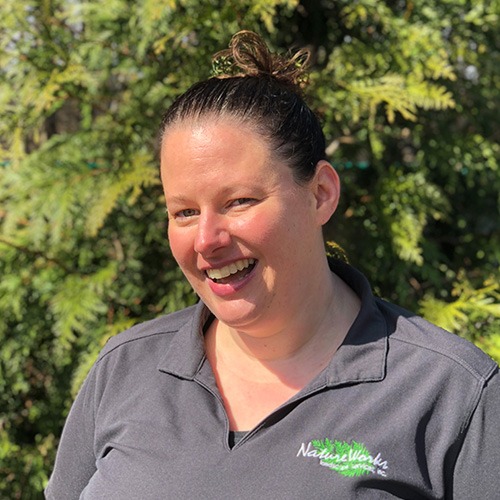 A person is smiling at the camera, wearing a dark polo shirt with a logo. The background shows greenery under bright sunlight.