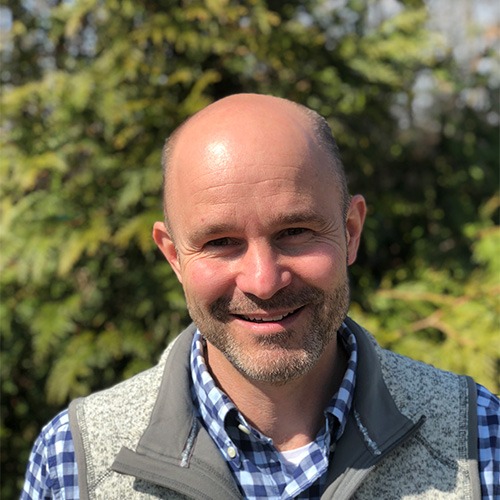 A person with a bald head and facial stubble is smiling outdoors. They're wearing a gingham shirt, grey vest, and trees in the background.