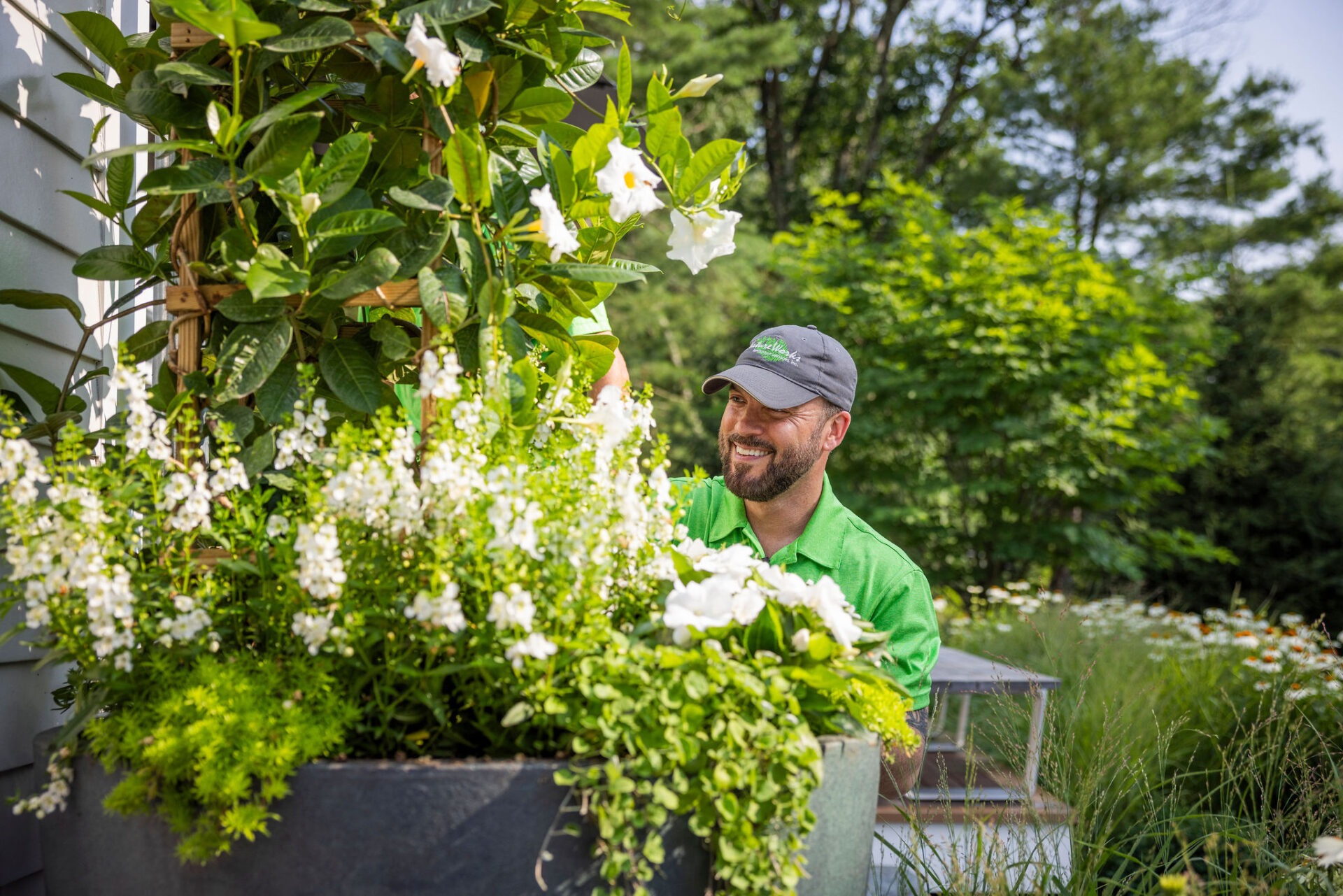 A person in a green shirt and cap cheerfully tends to a lush garden with vibrant white flowers, surrounded by greenery and sunlight.