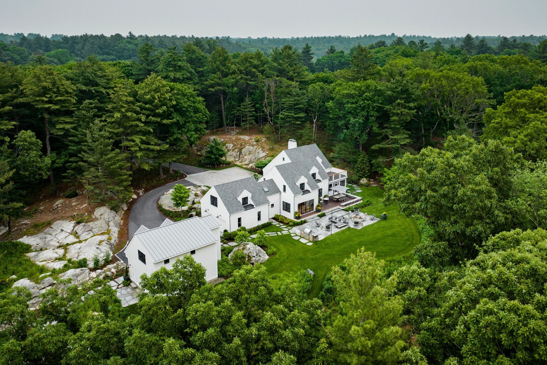 An aerial view of a large, white two-story house with multiple gables, surrounded by a green lawn, trees, and natural rock formations.