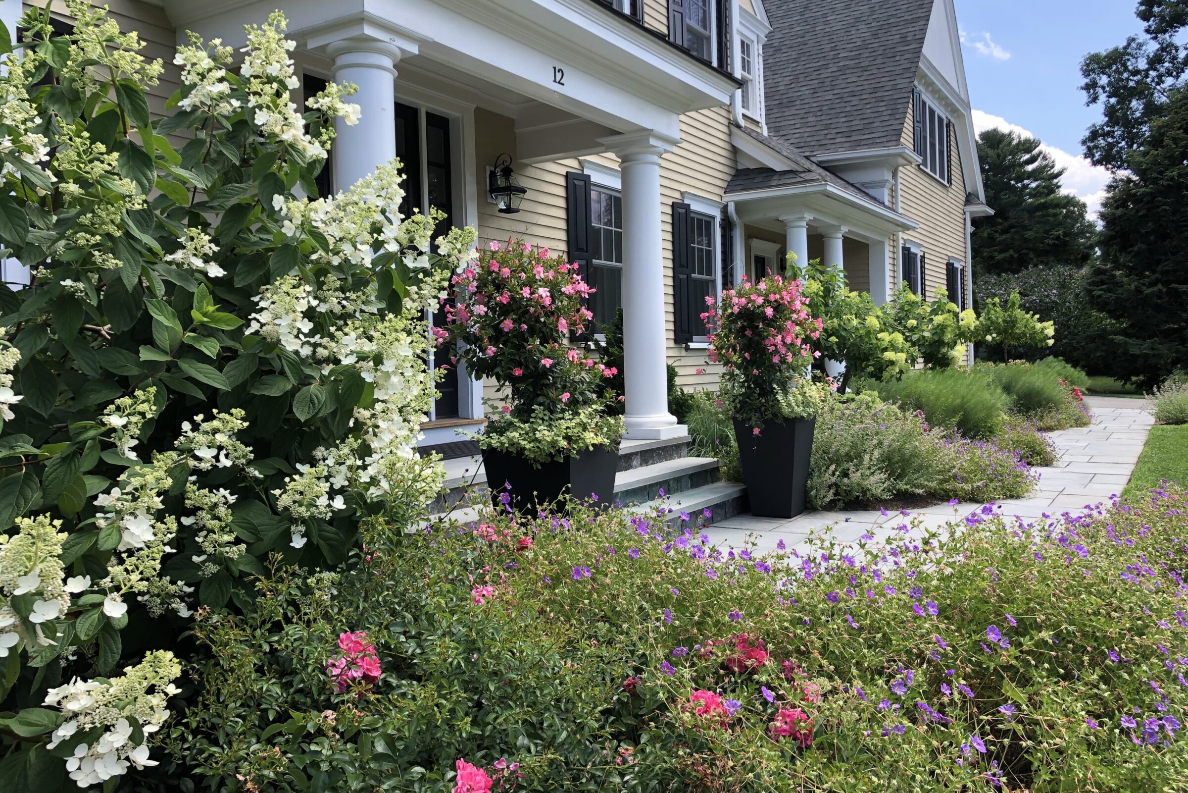 A well-maintained garden with vibrant flowers in front of a two-story house with classic architecture and substantial columns supporting the porch.