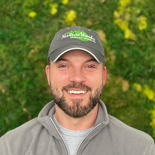 A smiling person wearing a baseball cap and a gray jacket stands before a blurred background with greenery and yellow flowers.