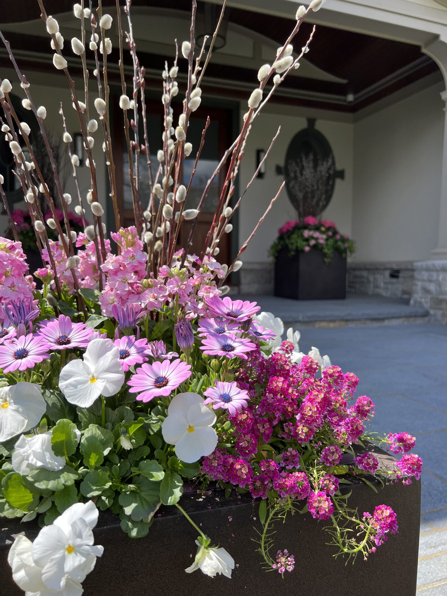 A vibrant flower arrangement with pink and white blossoms, displayed in a planter, with a blurred background featuring an outdoor porch and sculptural art.