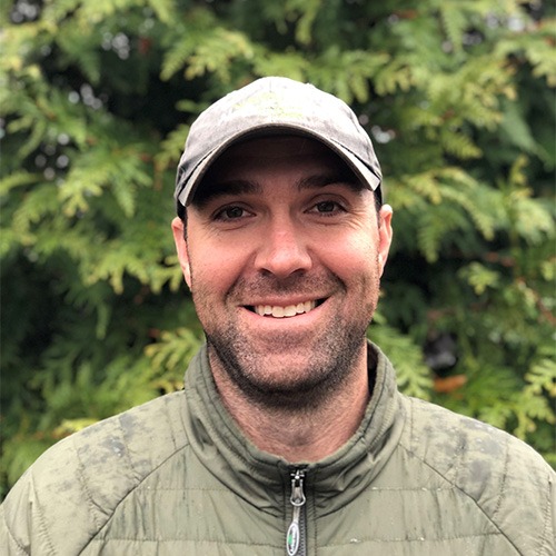 A person with a cap and a green quilted jacket smiles in front of bushy greenery. The outfit suggests cool weather, and the person appears content.