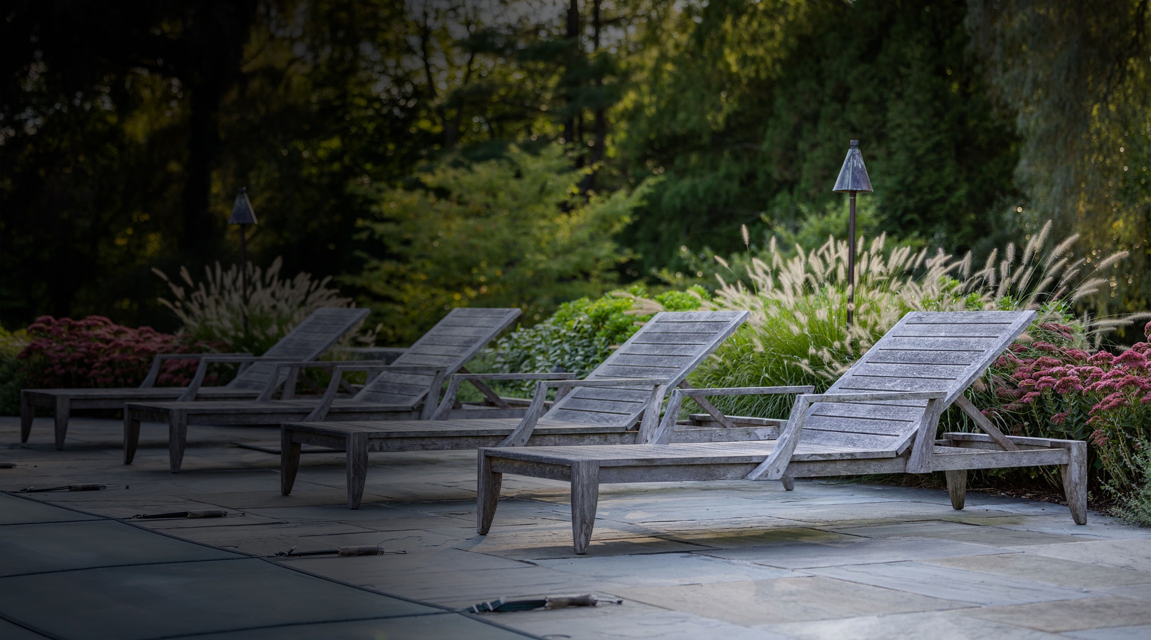 A serene garden setting with four empty wooden lounge chairs on a paved surface, surrounded by lush plants, tall grasses, and small lamps.