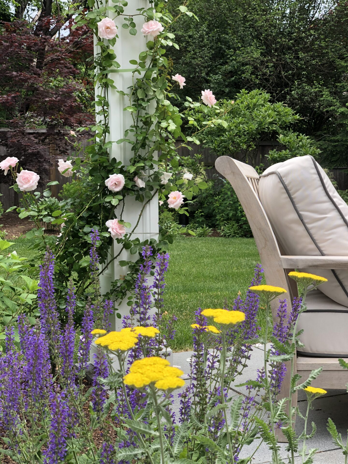 A serene garden with pink roses climbing a pillar, surrounded by purple and yellow flowers, next to a comfortable outdoor chair, suggesting relaxation.