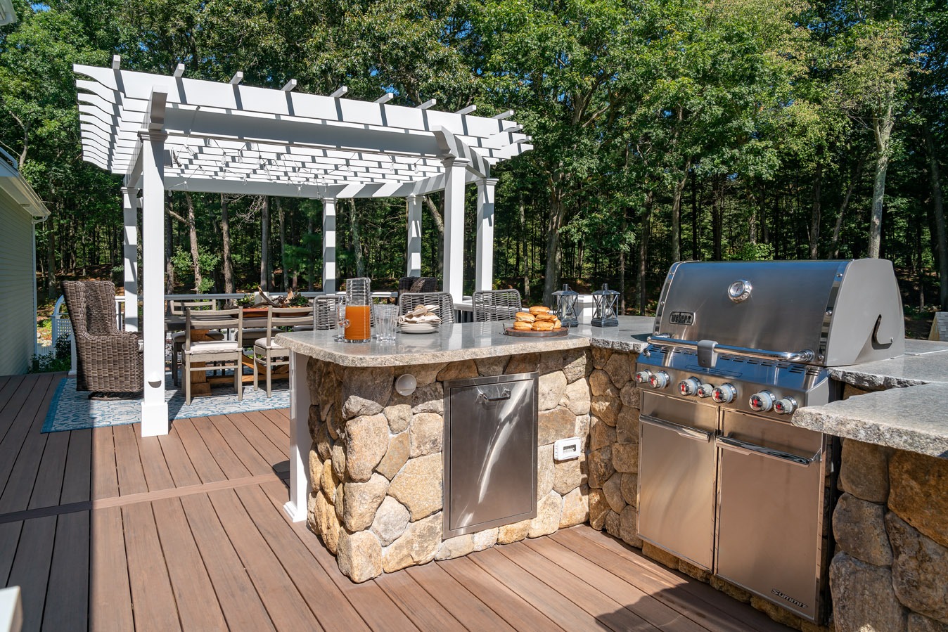 An outdoor kitchen with a stone counter, pergola, dining area, and grill on a wooden deck surrounded by trees on a sunny day.