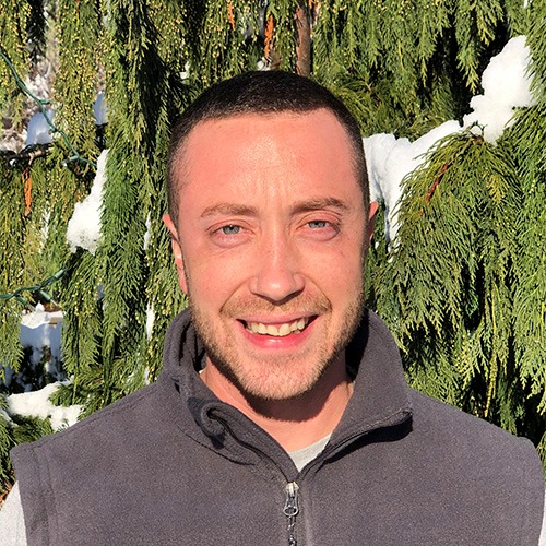 A person is smiling at the camera in front of snowy evergreen branches with sunlight on their face, wearing a gray zip-up top.