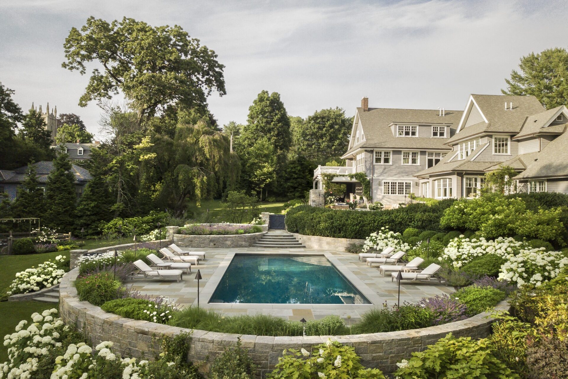 A luxurious estate with a large house, an outdoor pool surrounded by white flowers, sun loungers, lush trees, and a structured garden.