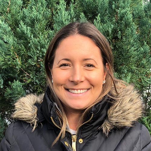 A smiling person with dark hair is wearing a black jacket with a fur-lined hood, standing in front of dense green foliage. Bright, casual, and outdoors.