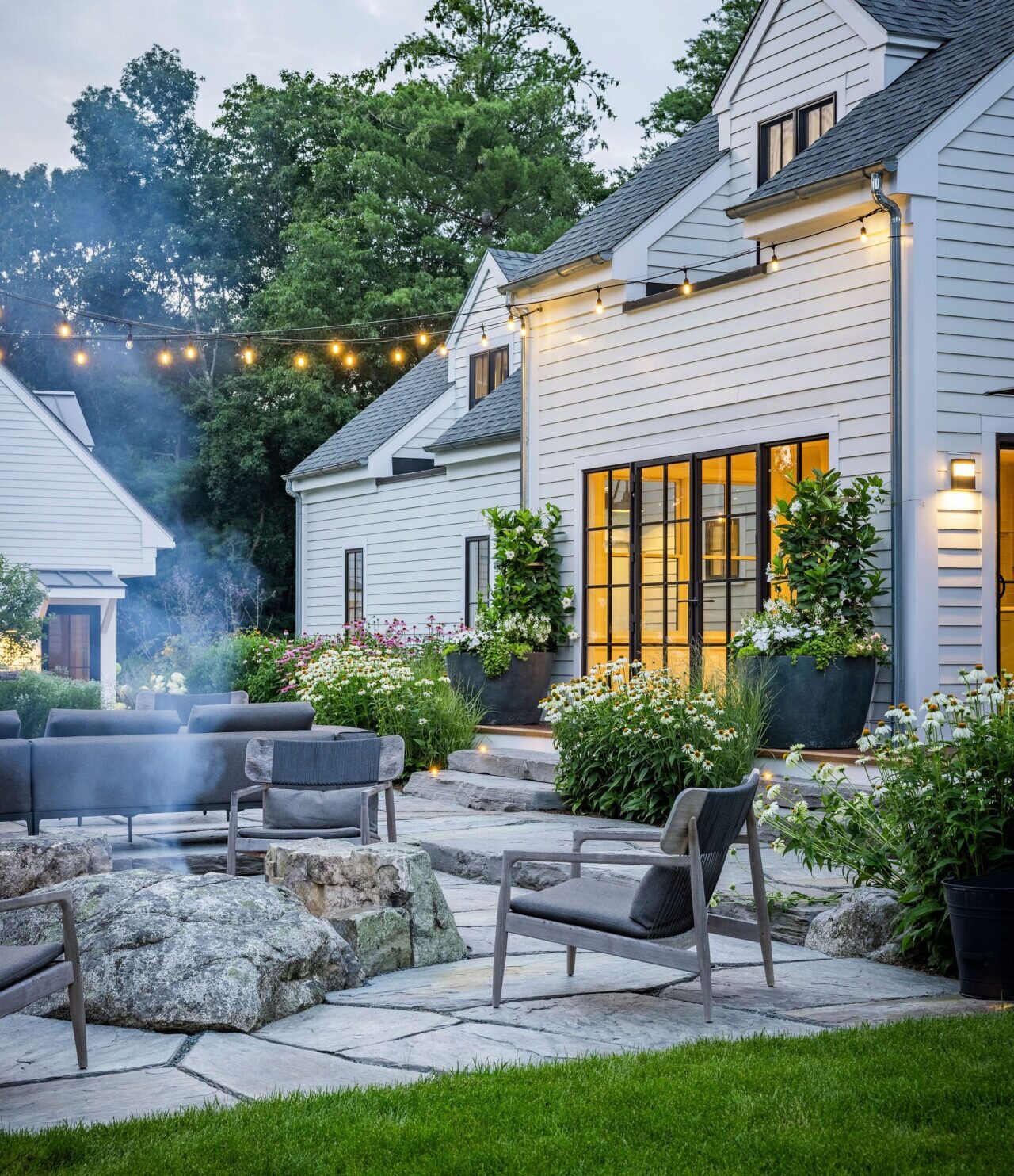 A cozy backyard setting at dusk with stylish outdoor furniture, string lights, plants, and a modern house with illumined windows and white siding.