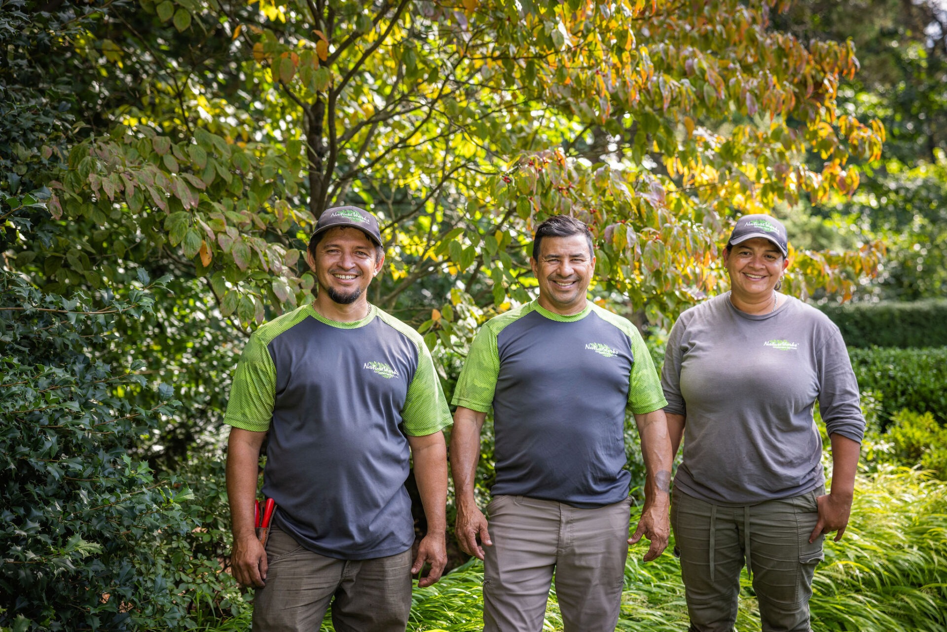 Three people in matching green work shirts stand smiling in a lush garden, exuding a sense of teamwork and professionalism amidst nature.