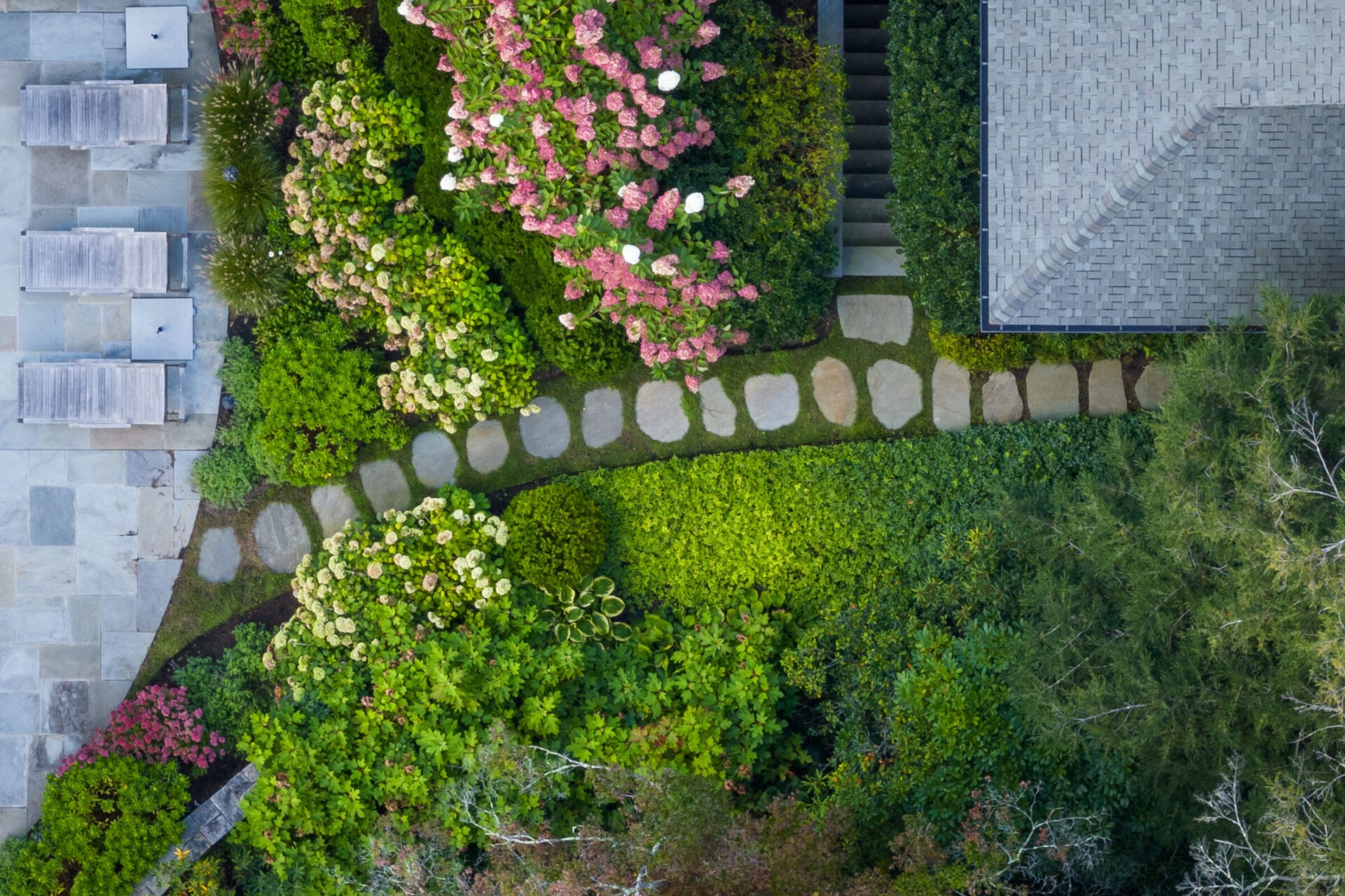 Aerial view of a landscaped garden with stepping stones, assorted plants, a staircase, and part of a building with a gray tile roof.