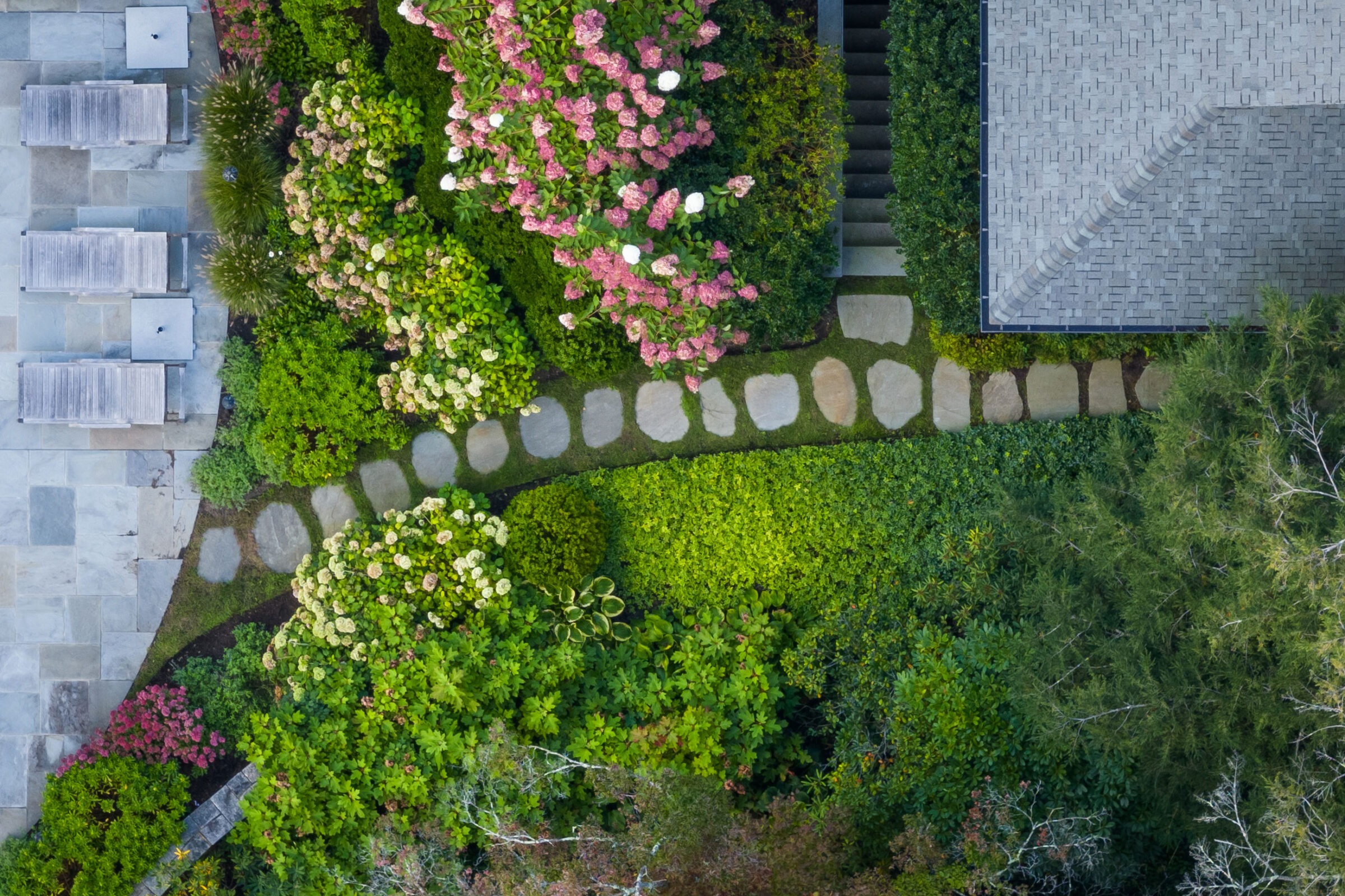 Aerial view of a landscaped garden with stepping stones, assorted plants, a staircase, and part of a building with a gray tile roof.