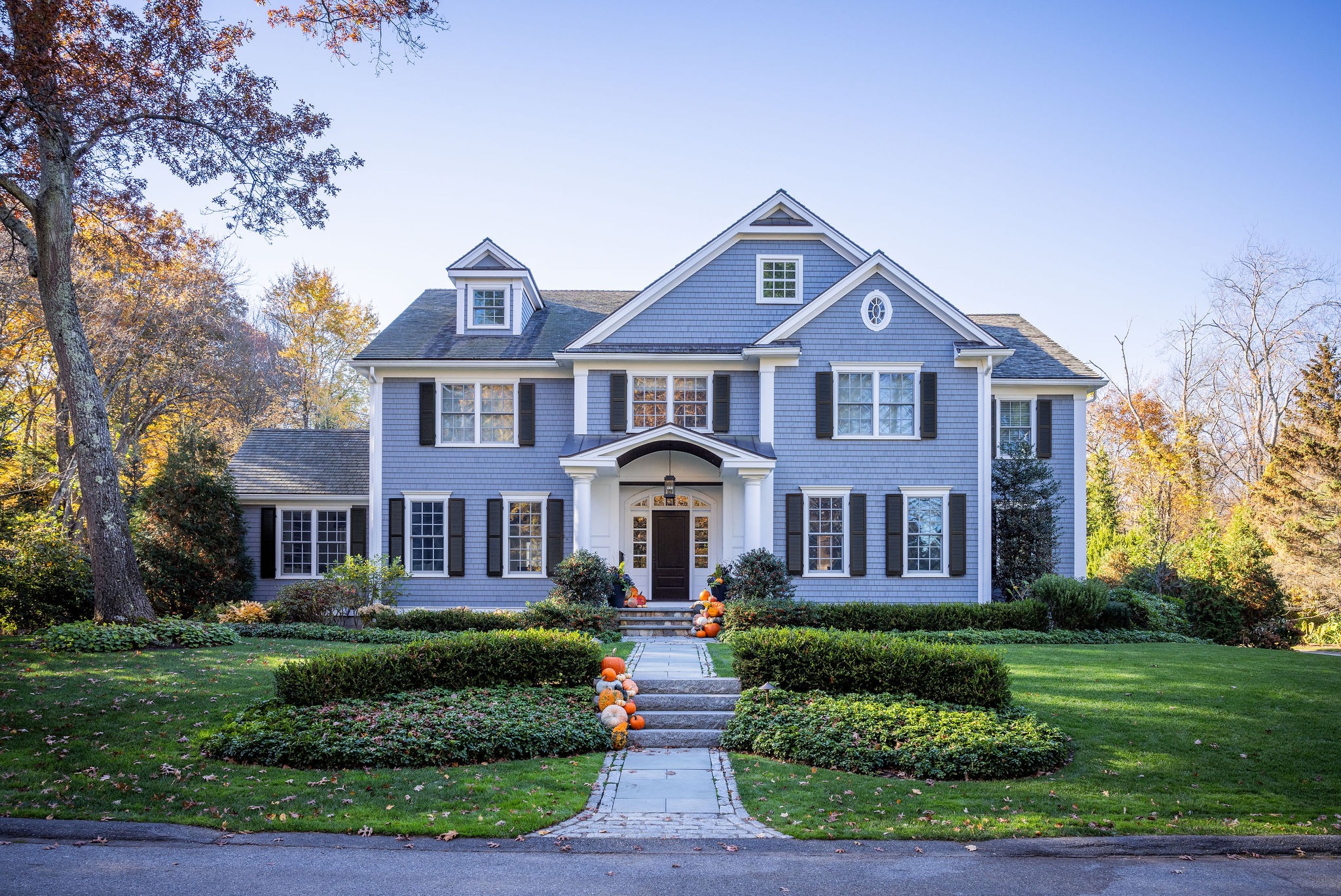 A stately blue two-story house with white trim and a covered entryway, surrounded by manicured hedges, a stone walkway, and autumnal trees.