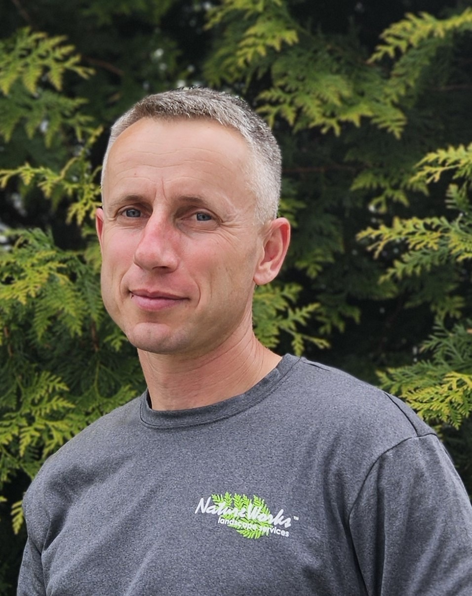 A person with short, graying hair and a slight smile stands outdoors wearing a grey T-shirt with a logo, against a blurred green foliage background.