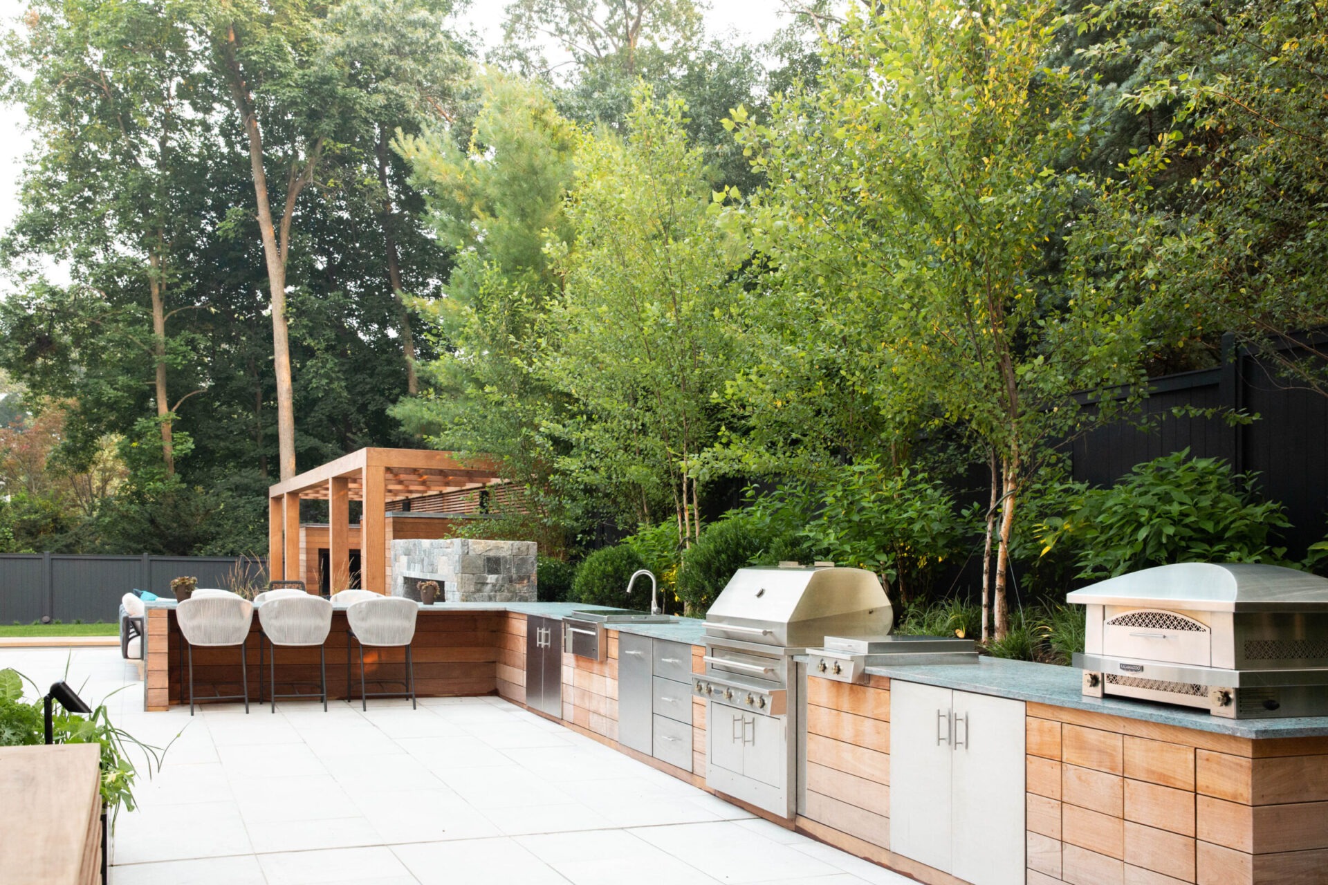 An outdoor kitchen with a pergola, modern appliances, and a bar seating area, set against a backdrop of green trees and a dark fence.