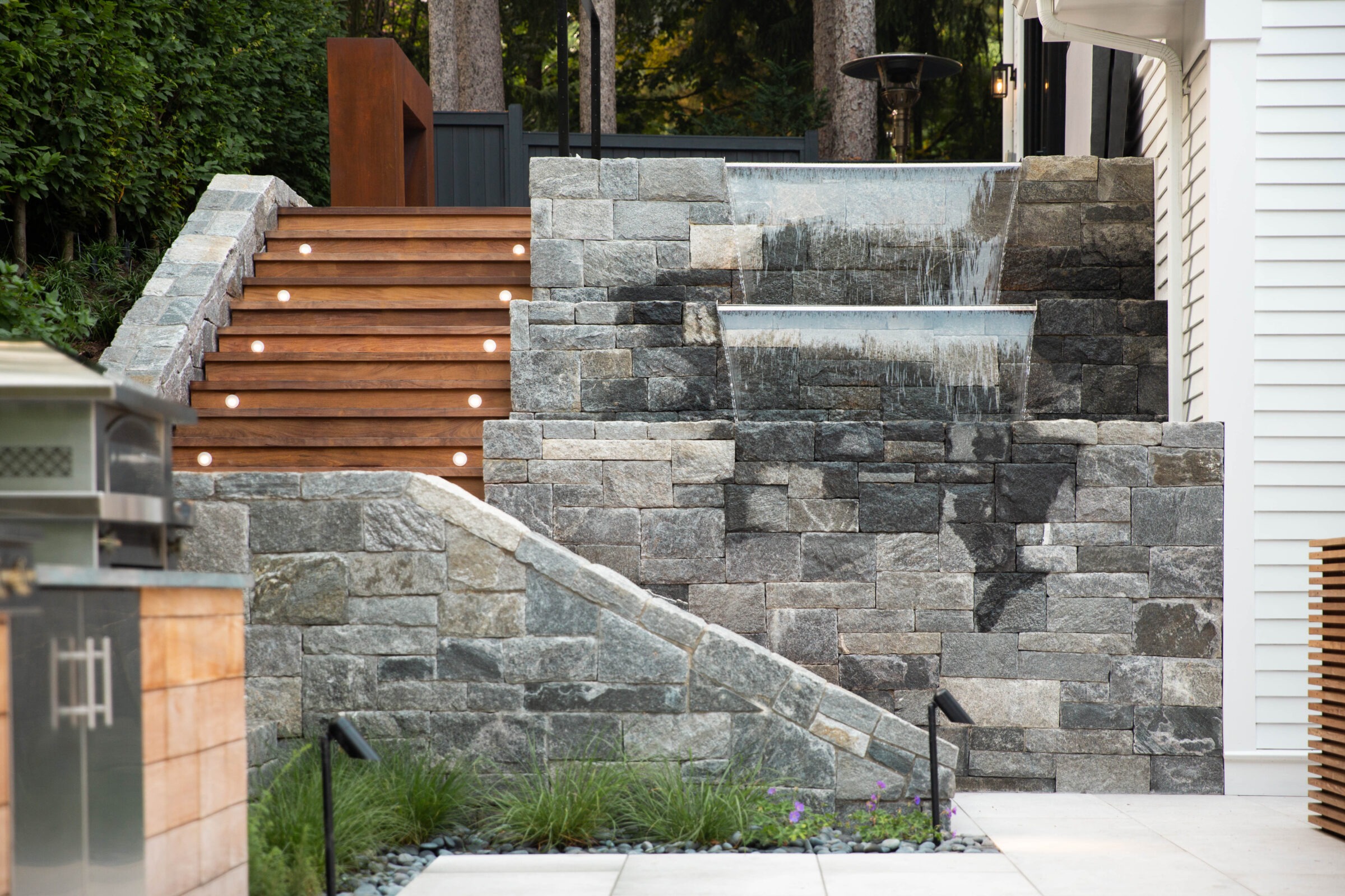 A well-lit outdoor staircase made of wood with built-in lights flanks a cascading stone water feature surrounded by landscaped greenery.