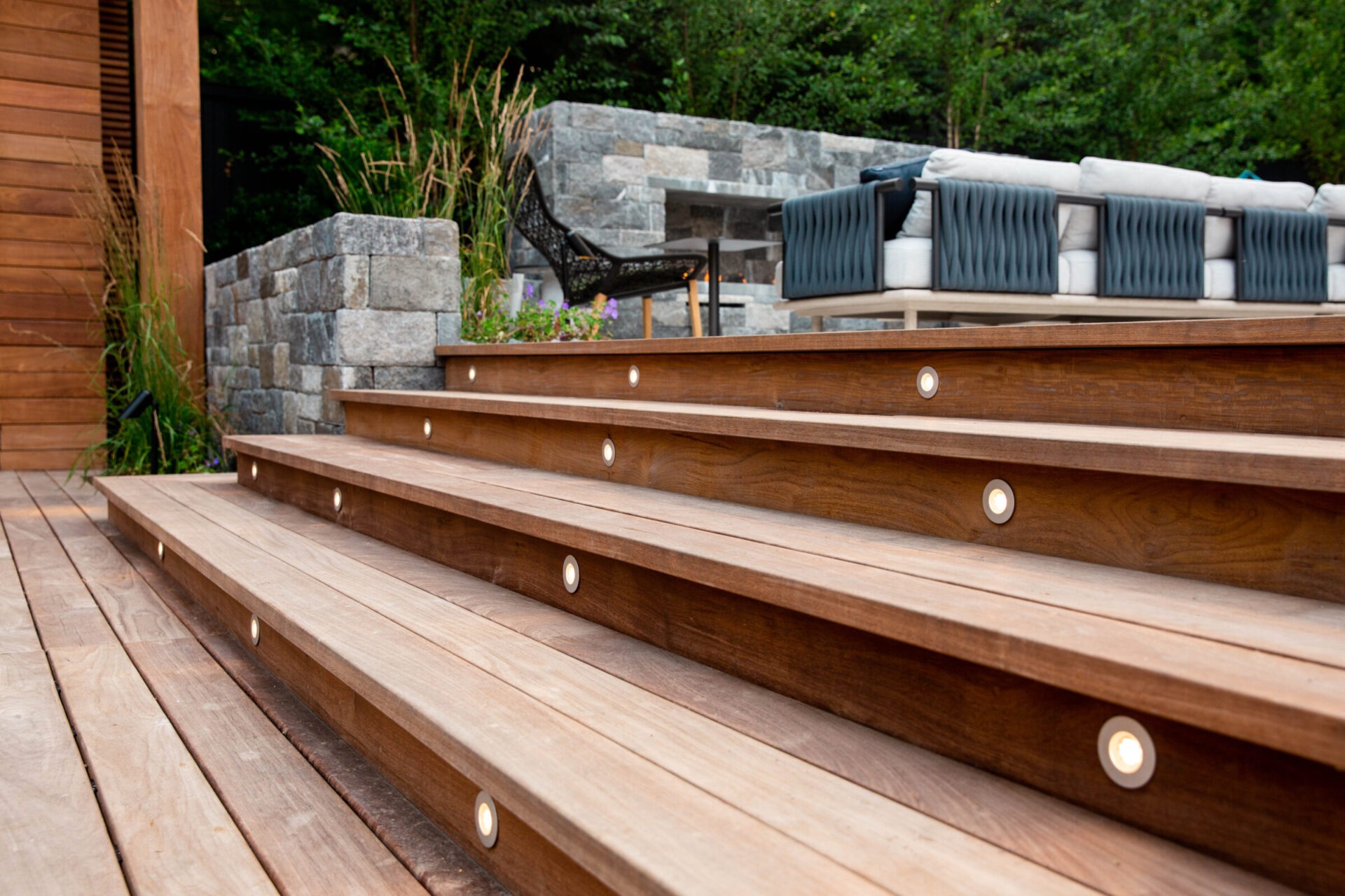 This image shows a well-maintained wooden deck with integrated lights on steps leading to a cozy outdoor seating area with cushions and a stone fireplace.