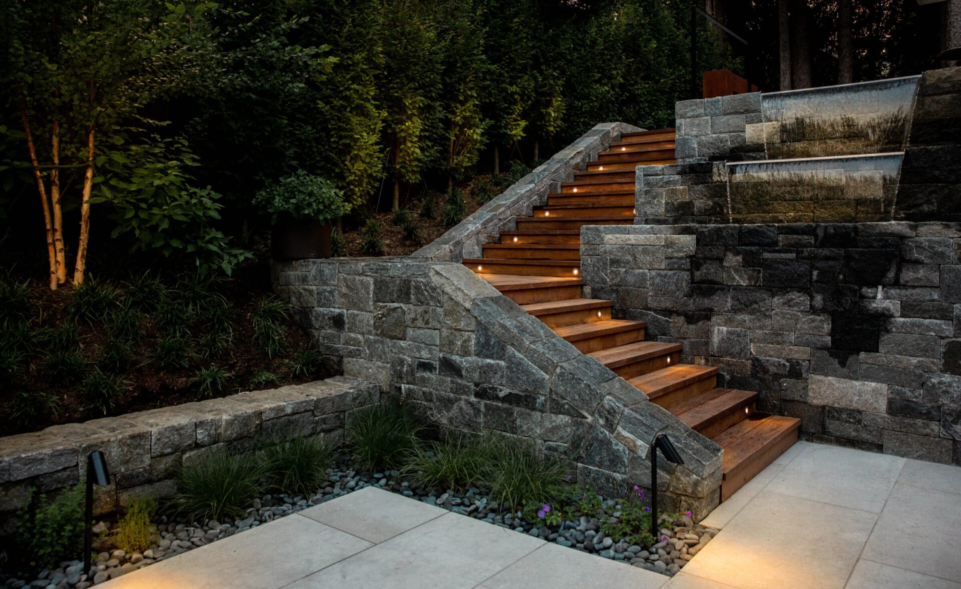 An outdoor staircase with wooden steps and stone walls, illuminated by embedded lights, surrounded by lush plants, and leading to a garden area.