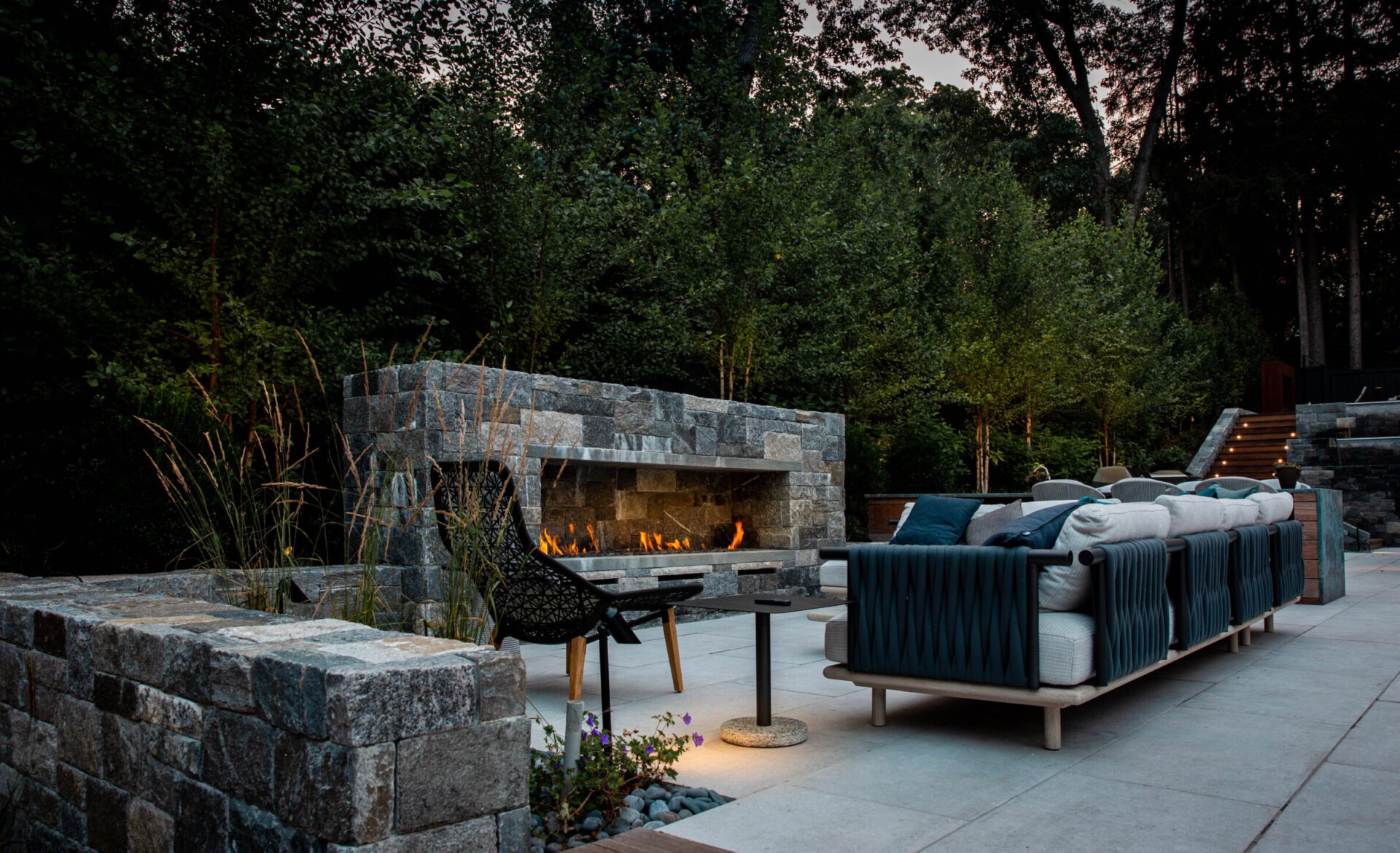An outdoor living area with a stone fireplace, comfortable seating, table, and surrounding greenery at dusk, exuding a cozy and inviting atmosphere.