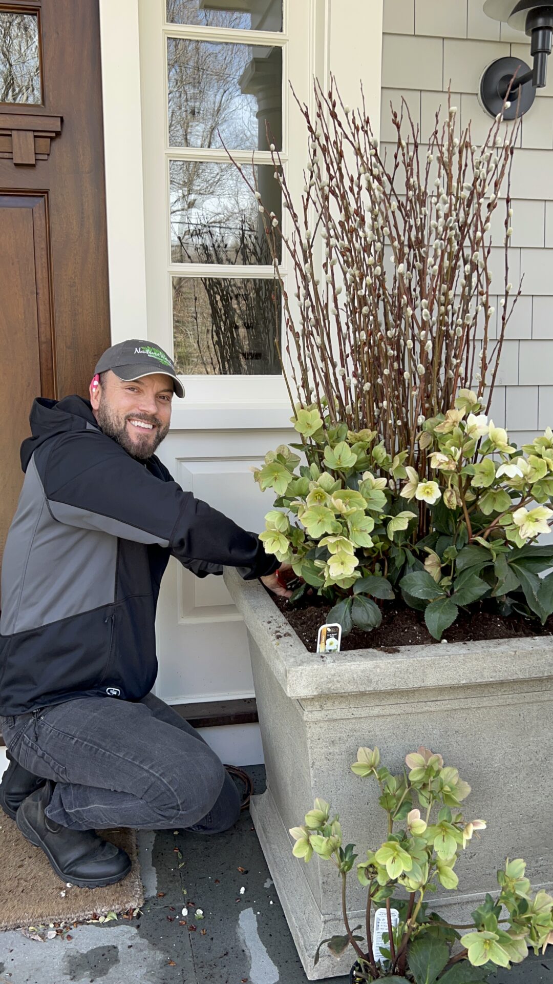 A smiling person is crouching next to a large planter with budding branches and blooming flowers outside a house, seemingly engaged in gardening.