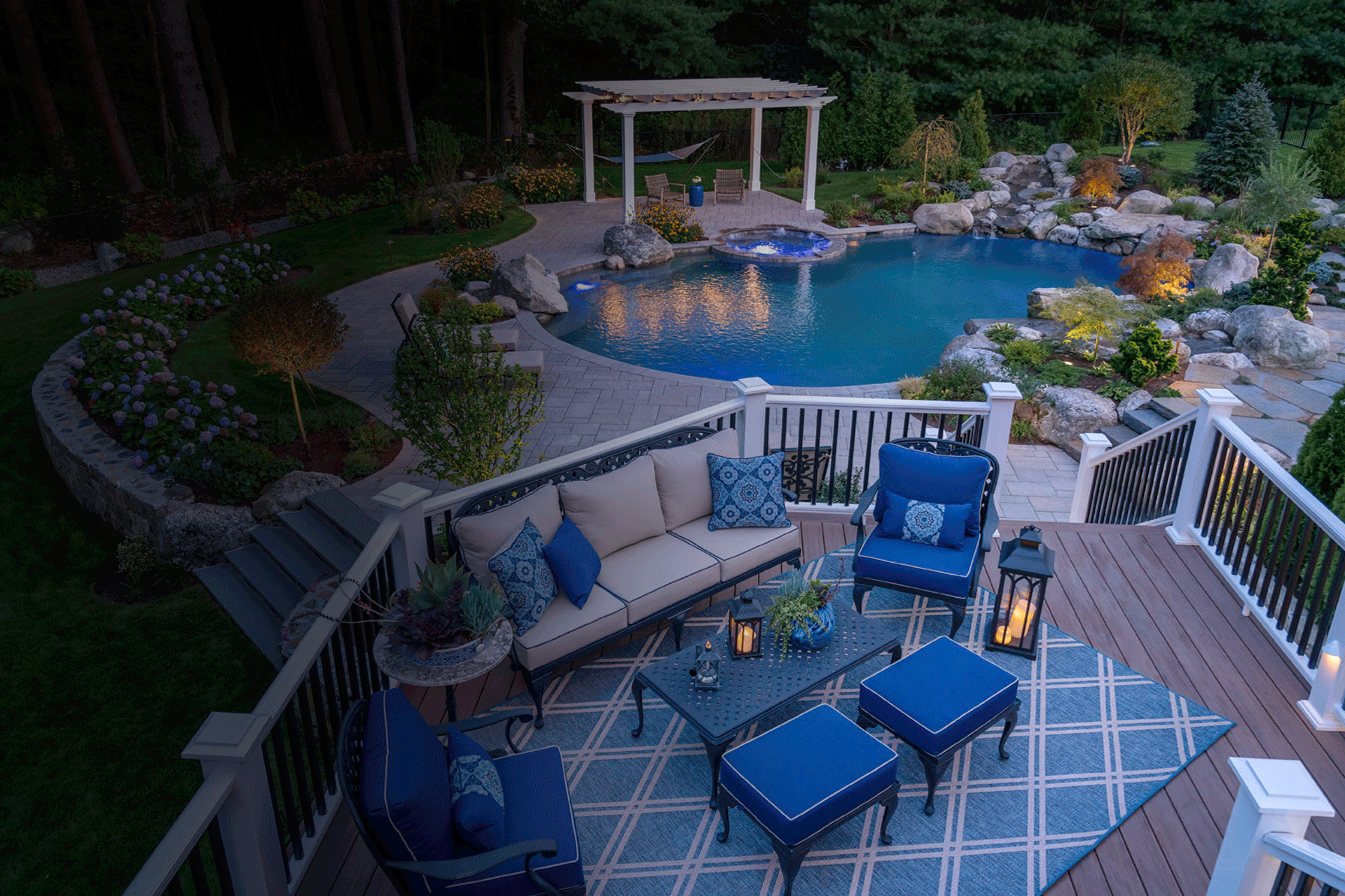 An elegantly furnished outdoor deck overlooking a beautiful swimming pool with blue cushions, flowers, rocks, and a pergola at dusk.