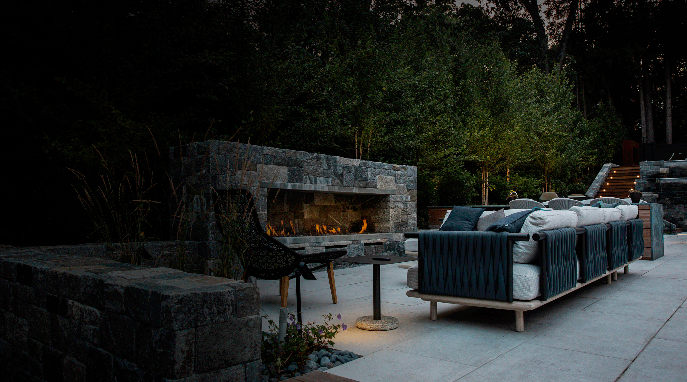 An outdoor patio area at dusk with a lit stone fireplace, comfortable seating, decorative pillows, and surrounding greenery giving a cozy ambiance.