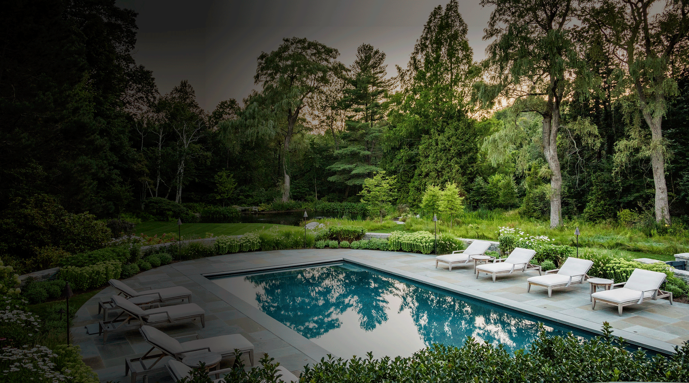 An outdoor swimming pool surrounded by lounge chairs, with reflecting trees, under a dusky sky nestled within a lush, landscaped garden.