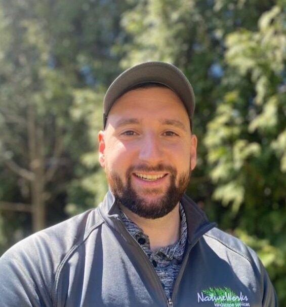 A person with a beard is smiling at the camera, wearing a cap and jacket, outdoors with green trees blurred in the background.