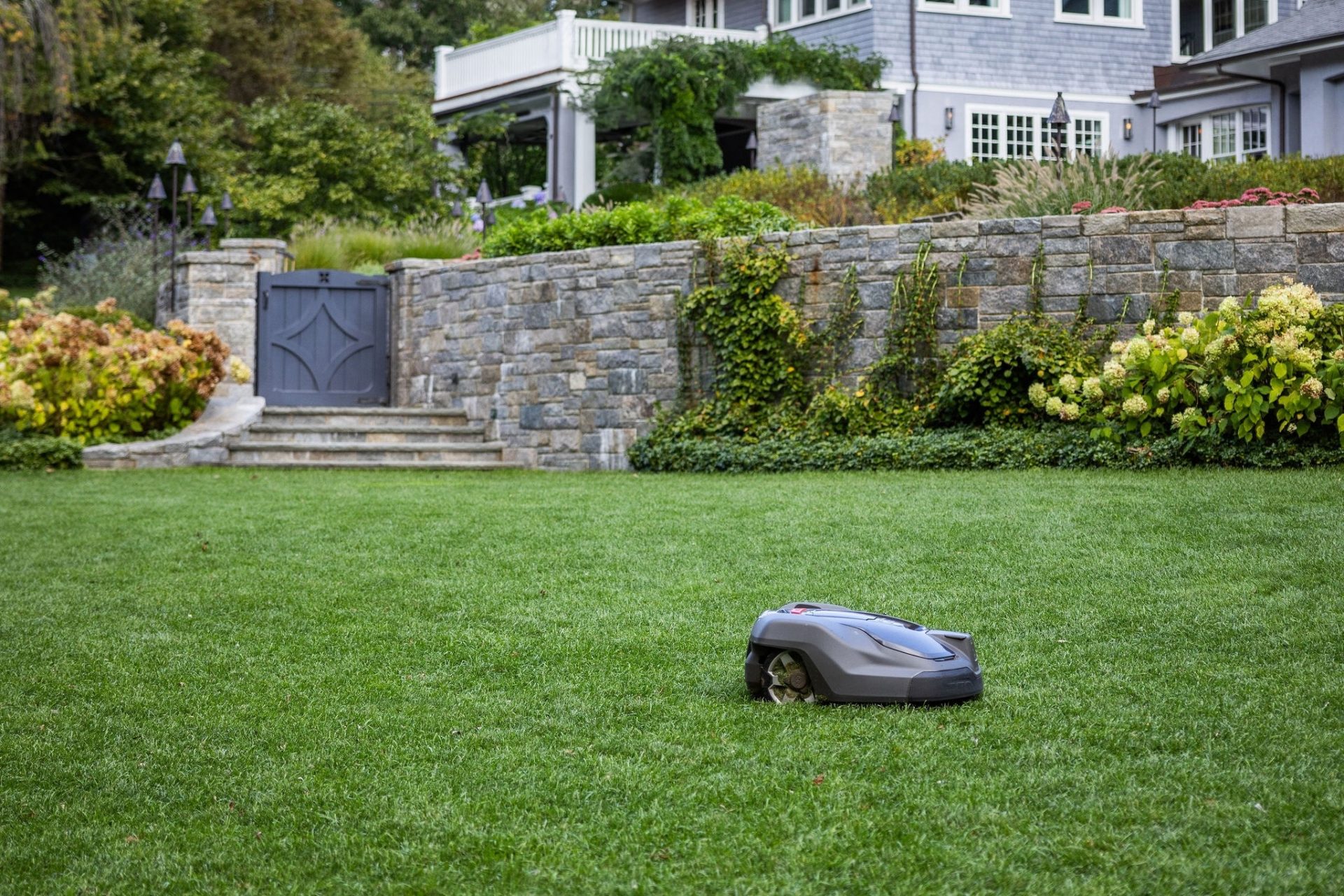A robotic lawn mower is trimming the grass in a well-manicured garden with a stone wall, greenery, and a glimpse of the house's exterior.
